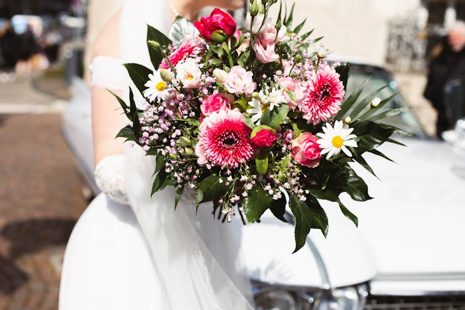 A bridal bouquet featuring a vibrant arrangement of pink and white flowers, including large pink gerbera daisies, delicate pink roses, and small white daisy-like blooms with yellow centers, all accent