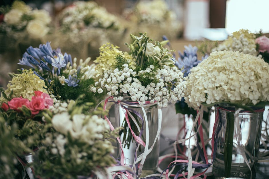 A collection of floral arrangements in glass vases, featuring predominantly white hydrangeas, with accents of pink roses, blue delphiniums, and small white baby's breath, all arranged in a natural, lo