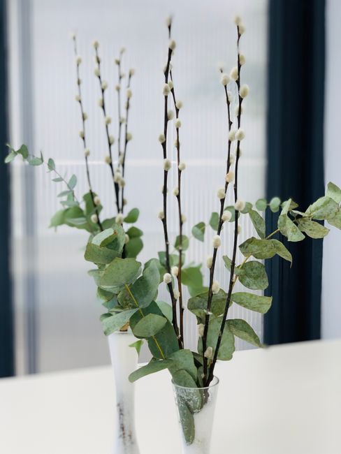 A clear glass vase containing a floral arrangement with tall, slender pussy willow branches adorned with soft, rounded catkins, complemented by sprigs of eucalyptus with oval-shaped green leaves. The 