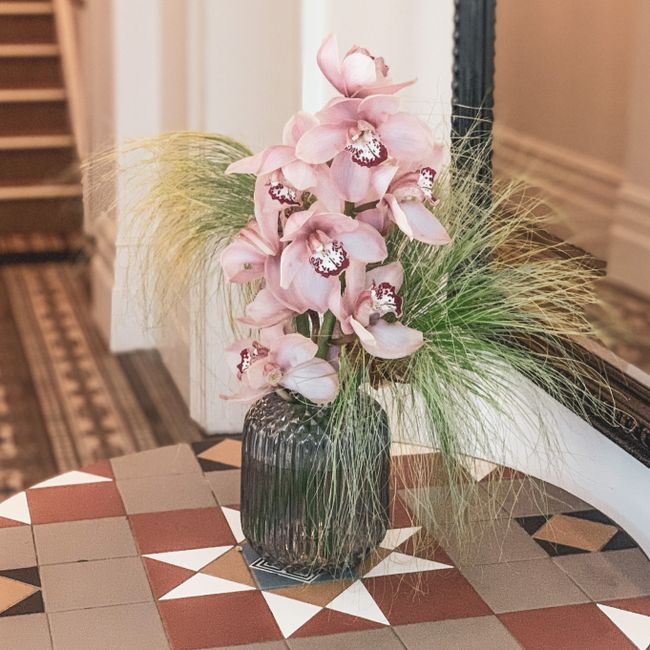 A floral arrangement featuring a tall bouquet of pink orchids with large, delicate petals and intricate dark markings on the lip, set in a textured black glass vase. The bouquet is complemented by wis